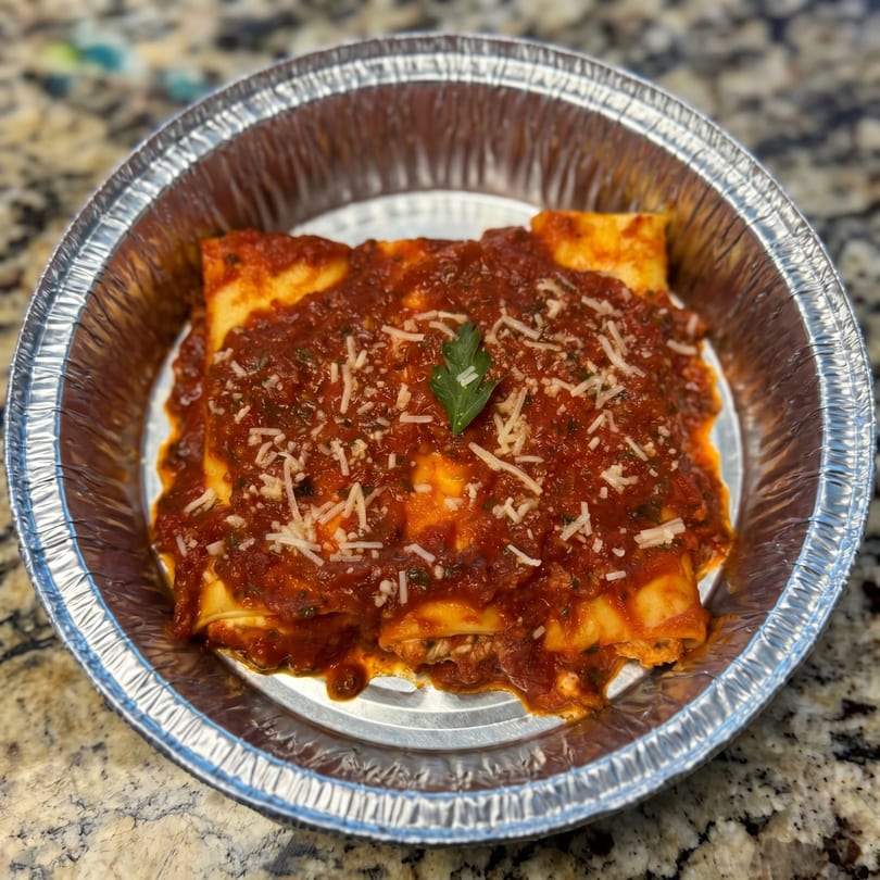 Baked cannelloni pasta with red sauce, parmesan cheese, and a parsley garnish in a foil pan.